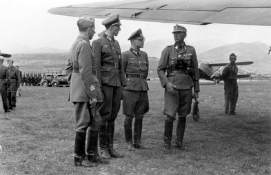 an Italian officer and three German officers in uniform standing beneath the wing of an aircraft on a grassed airfield